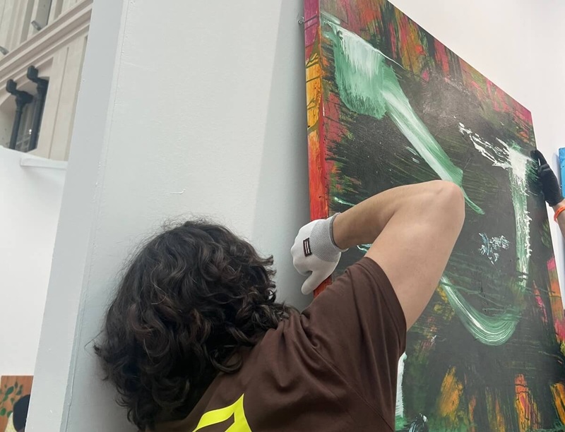 A logistics team member carefully installs a large abstract painting on a gallery wall, highlighting the behind-the-scenes work that supports art exhibitions.